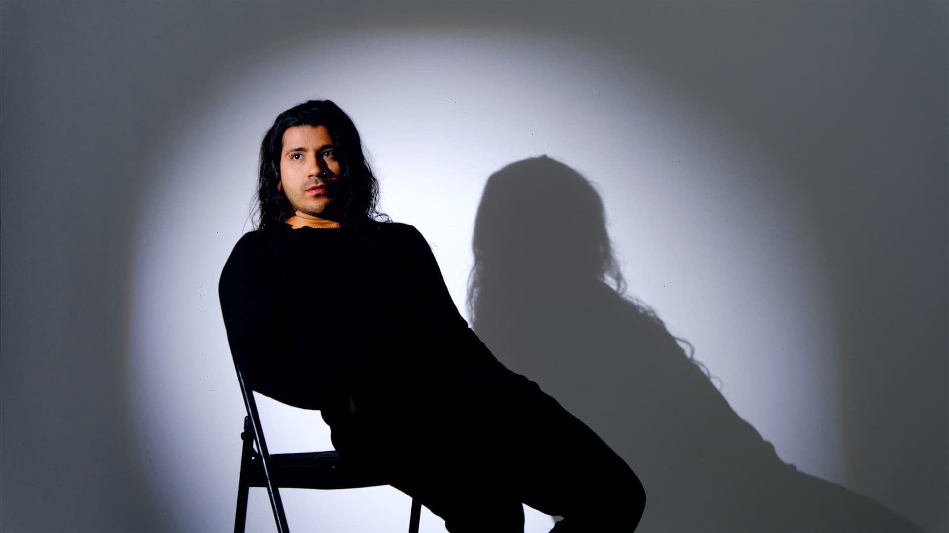 Claude D’Luv sentado en una silla, vestido de negro, con su sombra proyectada sobre una pared gris.