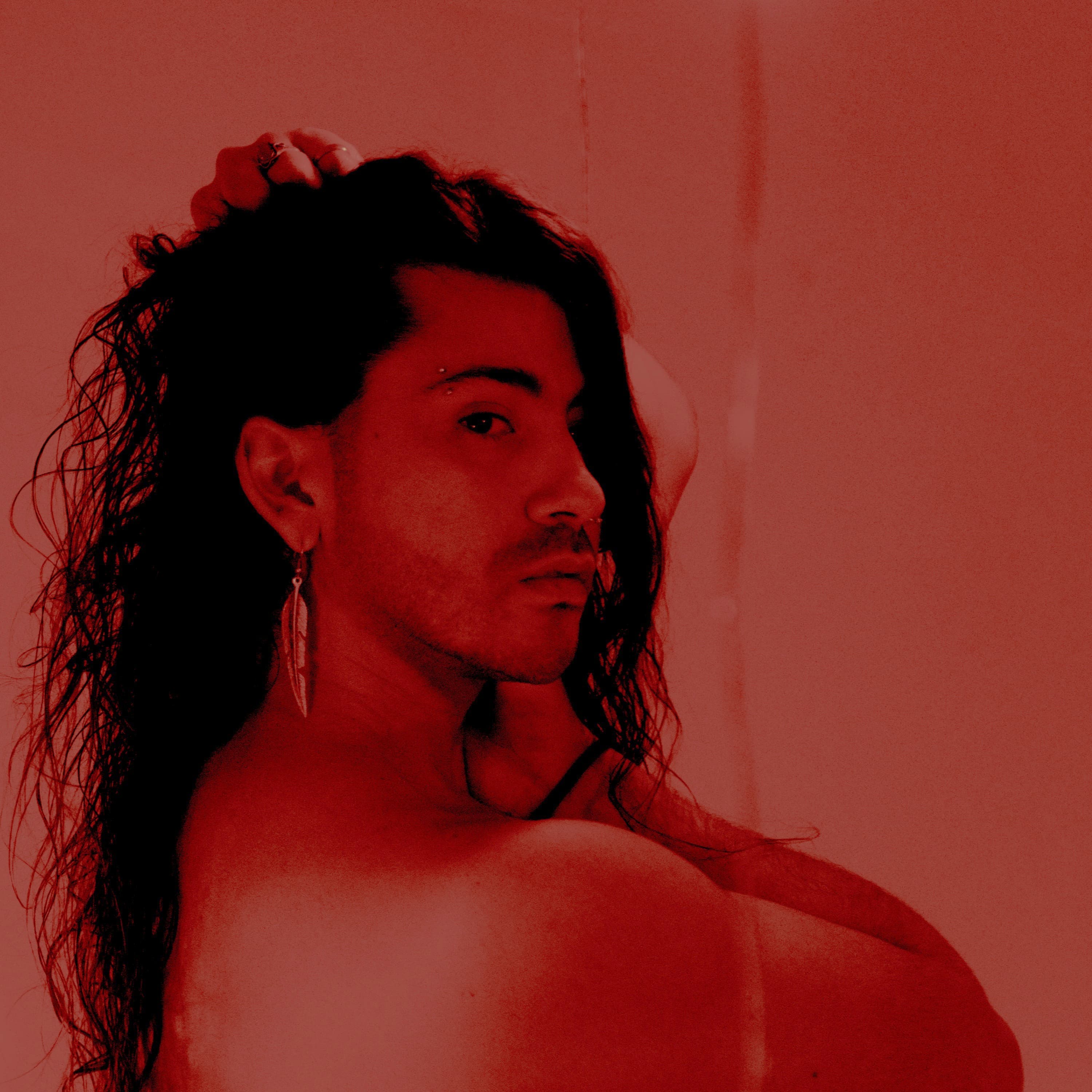 Retrato de Claude D’Luv con el cabello largo y una mano sobre la cabeza, iluminado en tonos rojos.